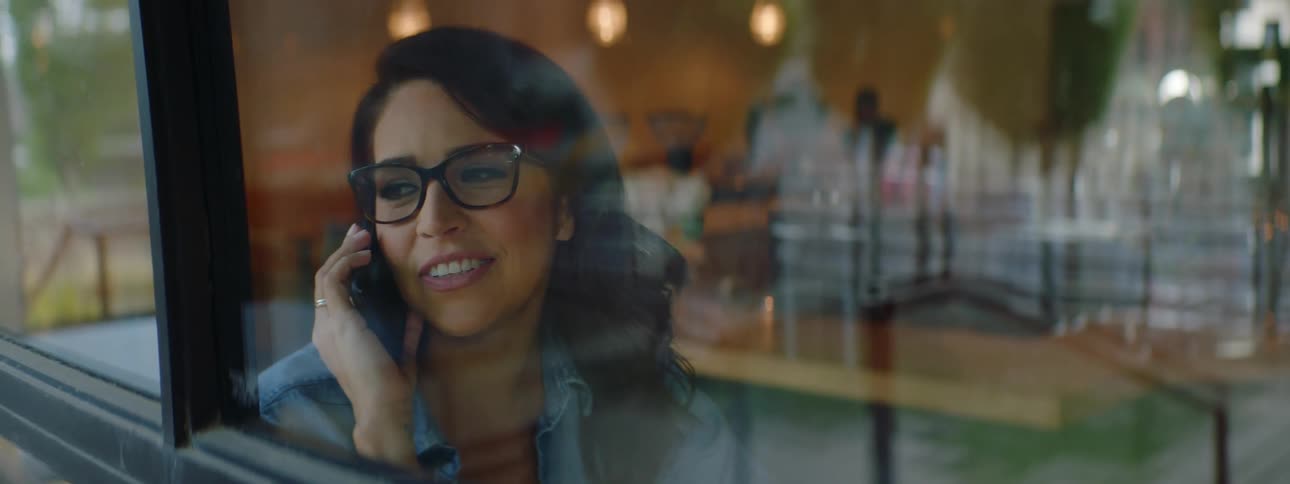 Young brunette woman wearing glasses talks on her smartphone while sitting next to a window in a restaurant