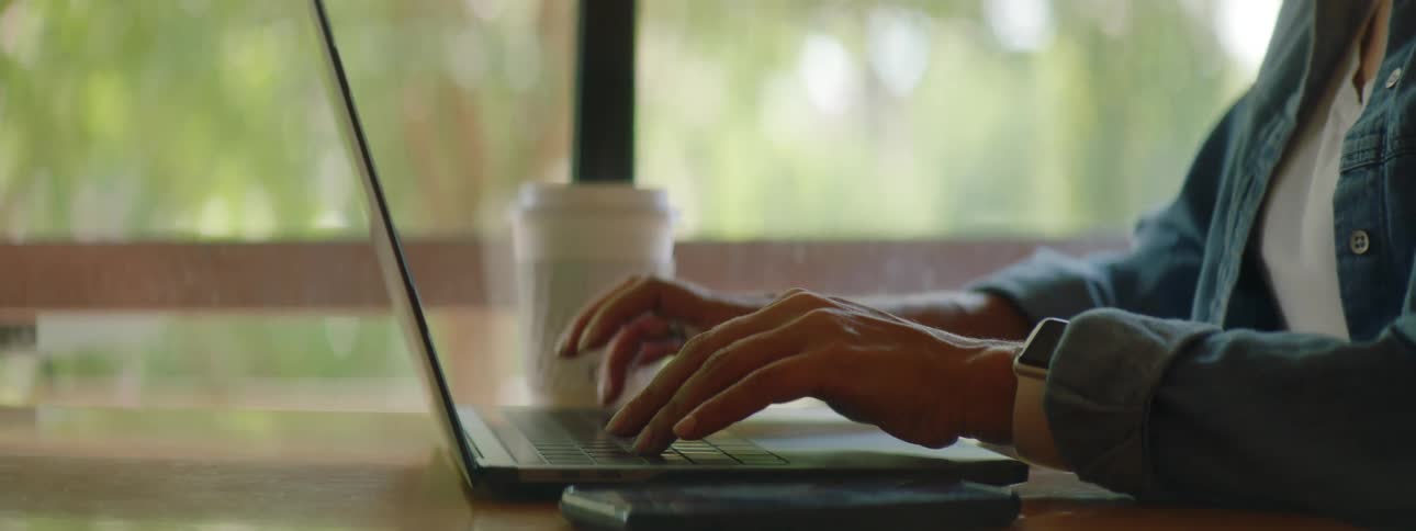 A woman types on her laptop while sitting at a table near a large window