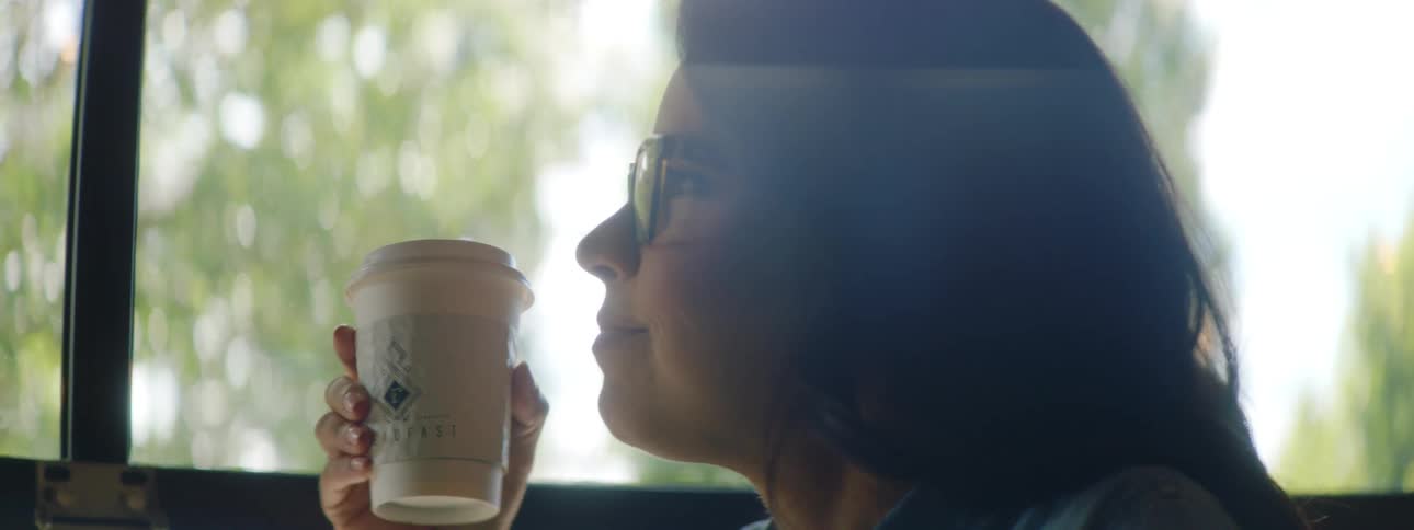 A woman sitting at a table with her laptop takes a sip from a disposable coffee cup