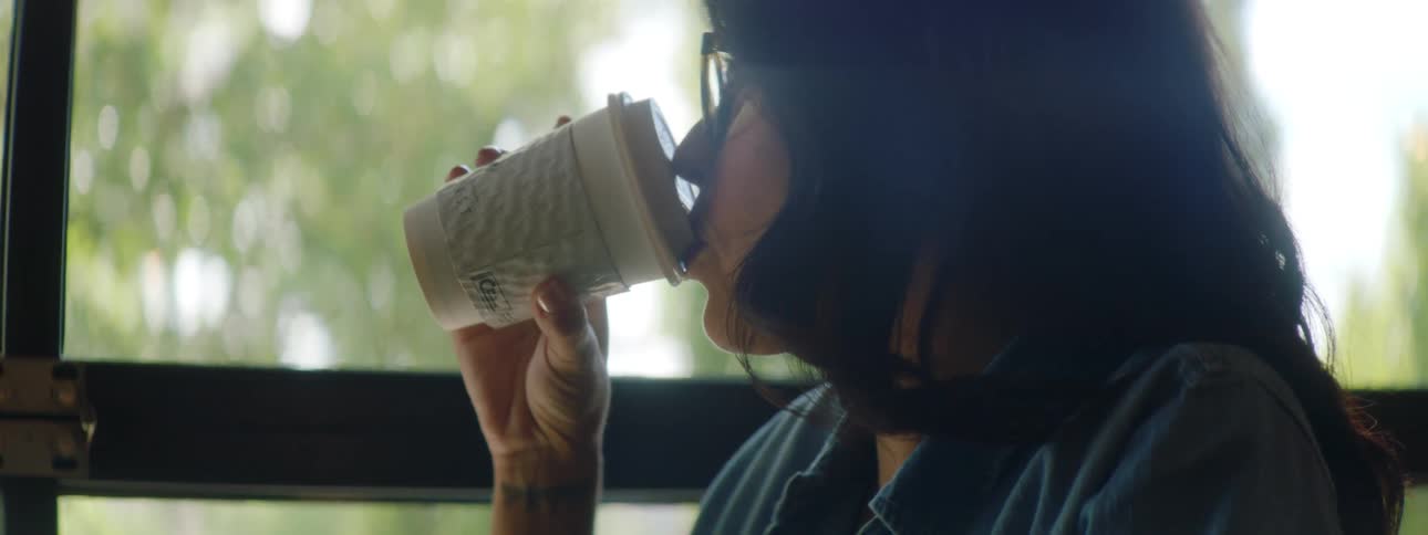 A woman typing on a laptop while sitting at a table stops to take a sip from a disposable coffee cup