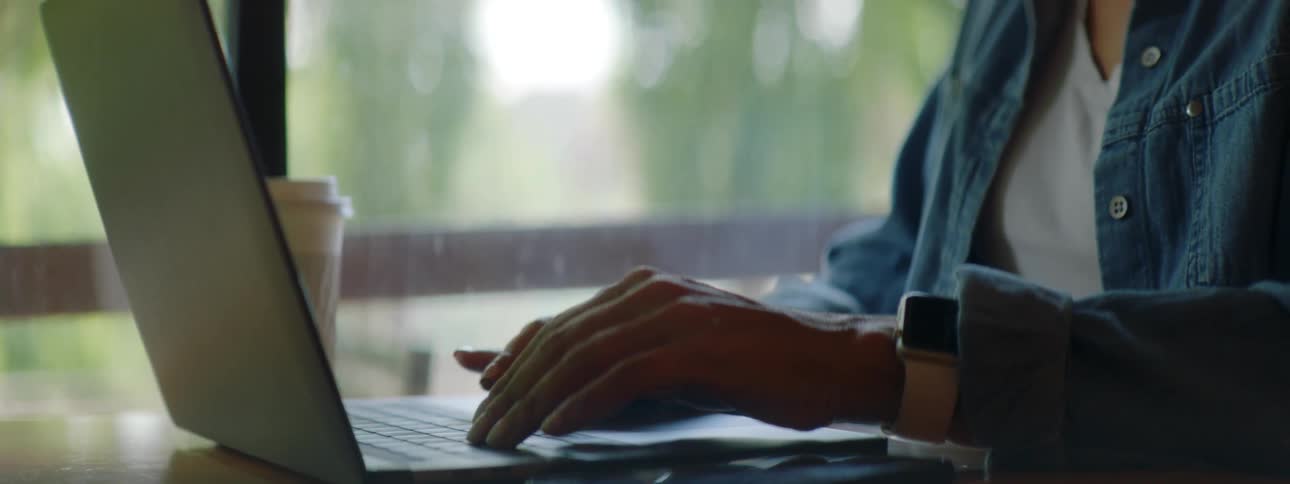 Brunette woman with wavy hair a blue shirt and glasses sits at a table and uses a grey laptop in a cafe