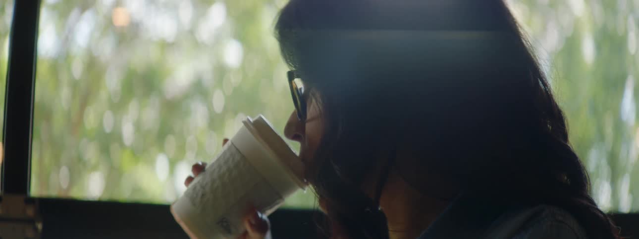 Woman with wavy hair a blue shirt and glasses sits at a table uses a laptop while drinking coffee at cafe