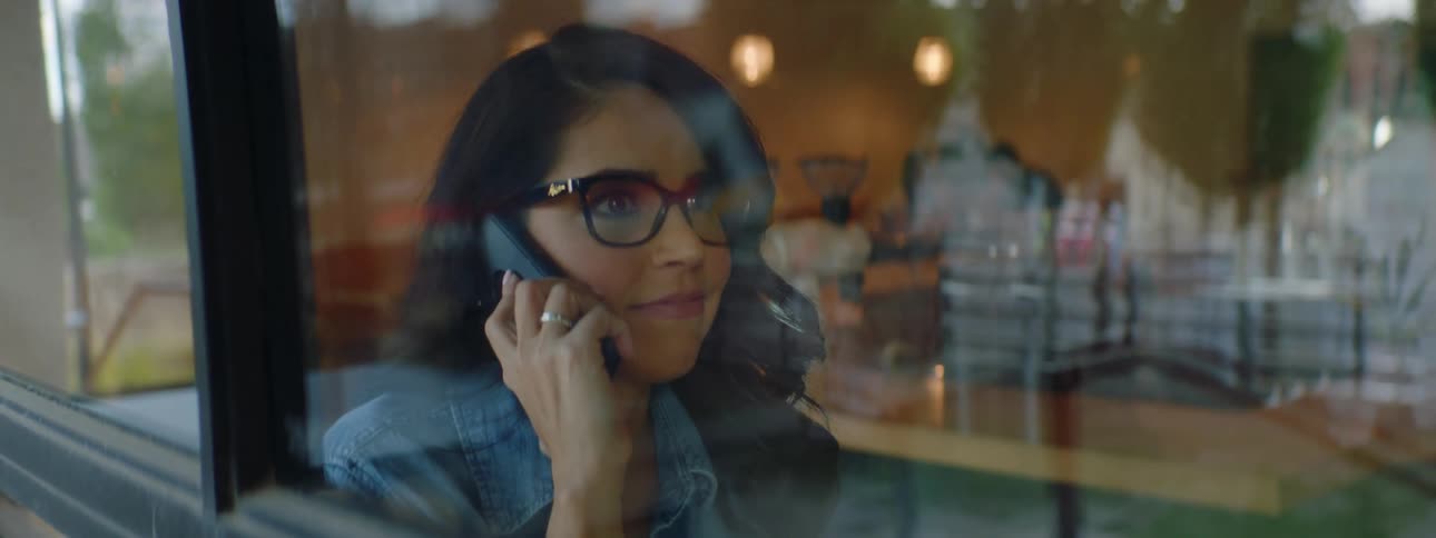 A woman sitting next to the window inside of an eatery happily answers her cell phone