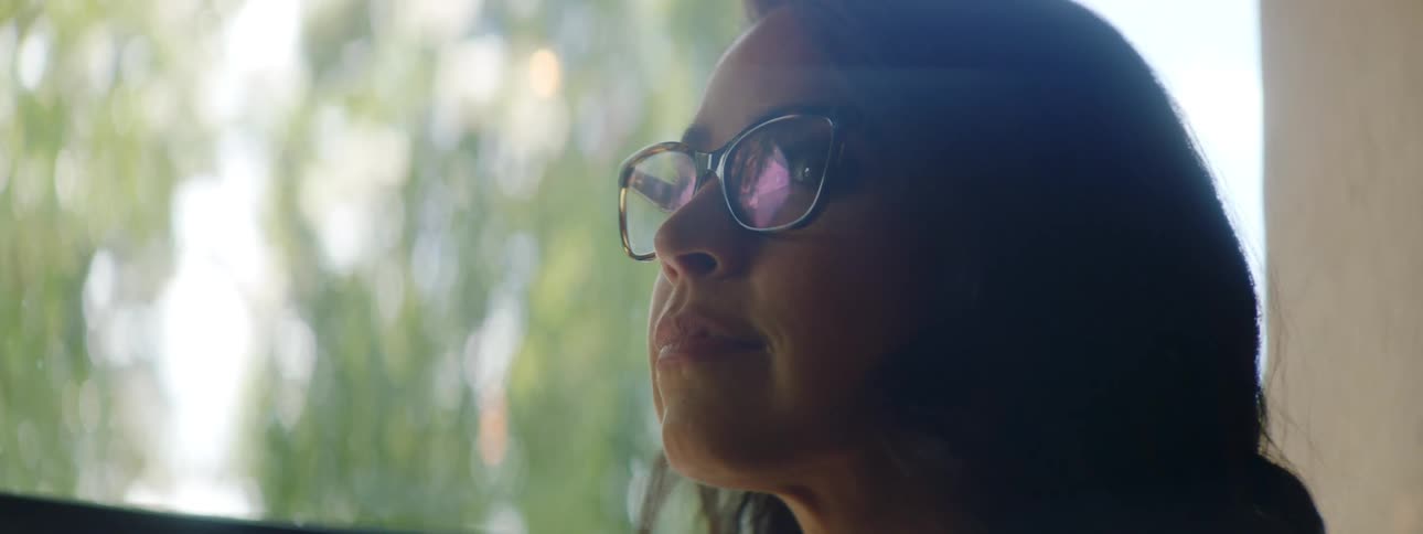 A woman typing on a laptop raises her head to look out the window before slowly turning to smile into the camera
