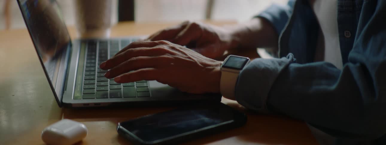 Brunette woman with wavy hair glasses and a blue shirt sits at a table and uses a grey laptop in a cafe