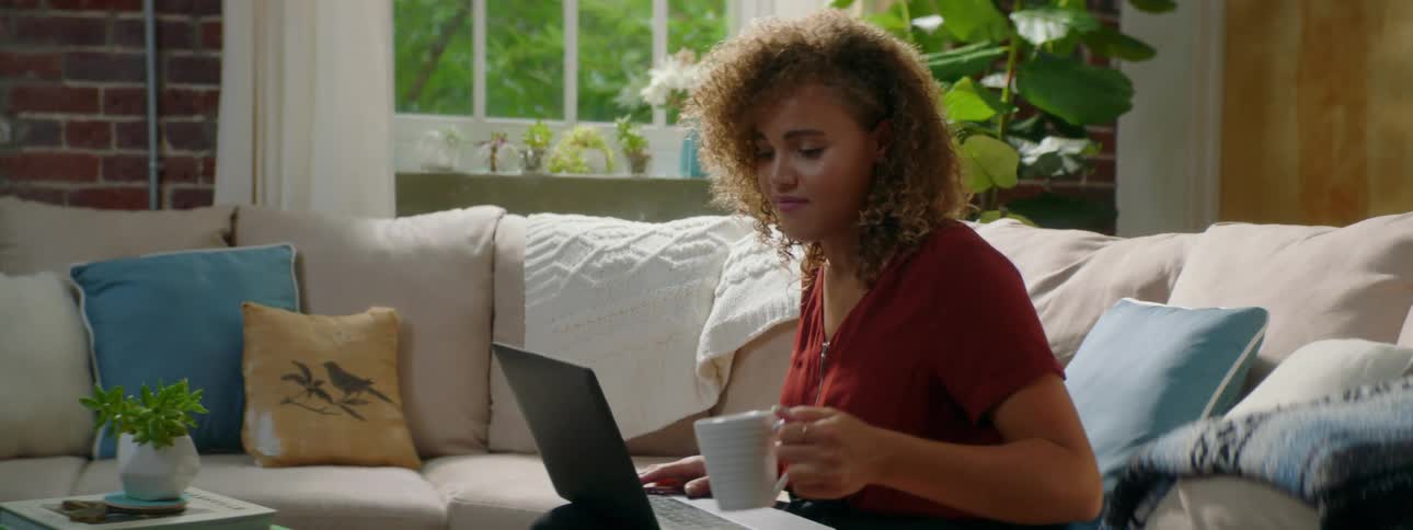 Woman with red curly hair and a red blouse drinks a cup of coffee while using a laptop in the living room