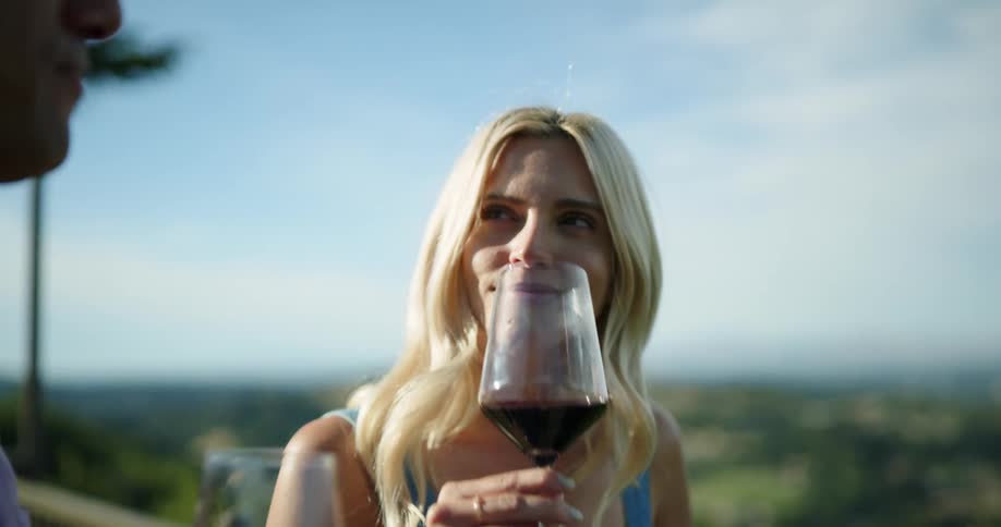 A young woman standing on a balcony overlooking the countryside sips wine while talking with a man