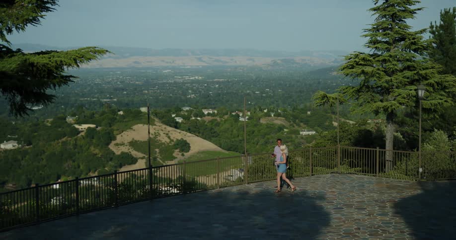 A man and woman carrying glasses of wine approach the guard rail of a lookout and drink while gazing out at the vast countryside below