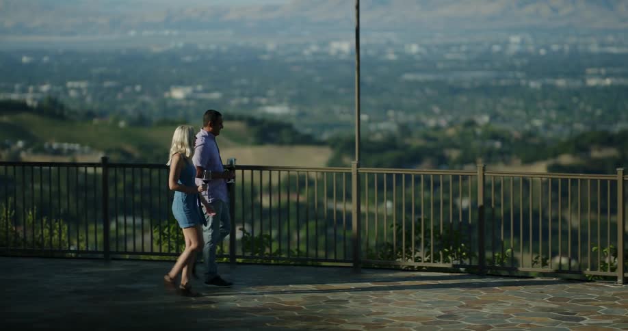 A man and woman on a date stroll along a walkway carrying wine stopping at an elevated lookout point