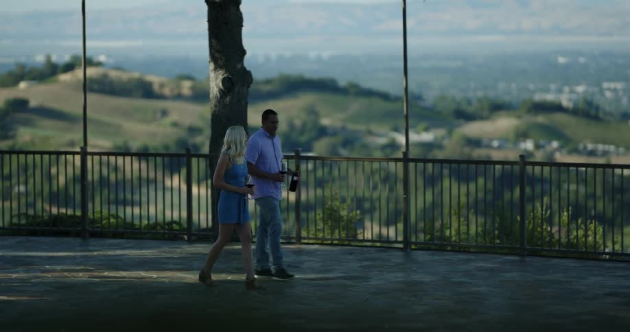 A man and woman walking along with glasses of wine and the bottle with the city skyline in the background