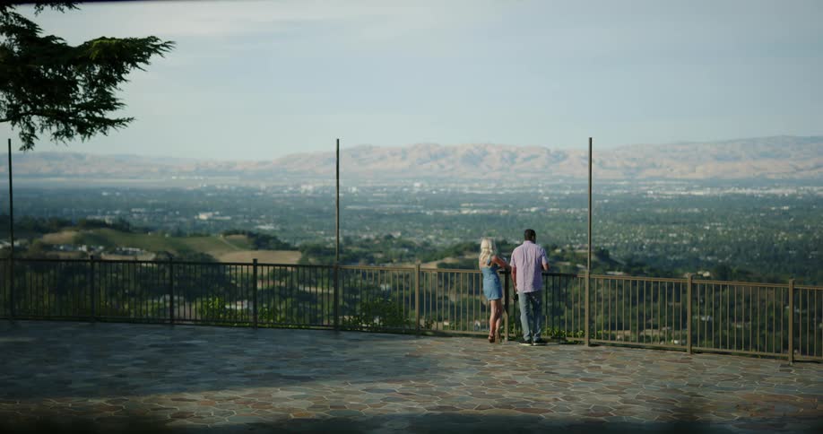 A man and woman stand against a railing and look out at the landscape while drinking wine