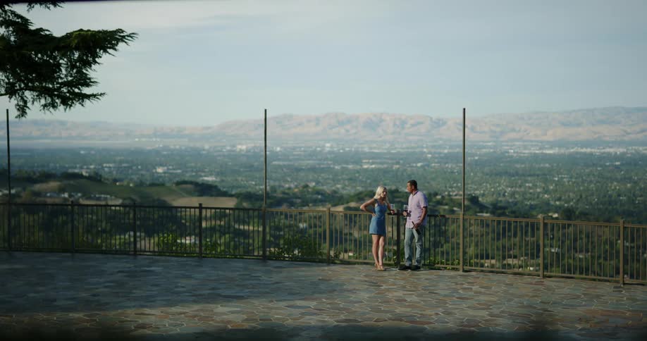 A man and woman stand on a terrace with the city skyline behind them and talk