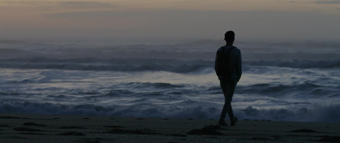A man walks across the sand toward the waves at sunset