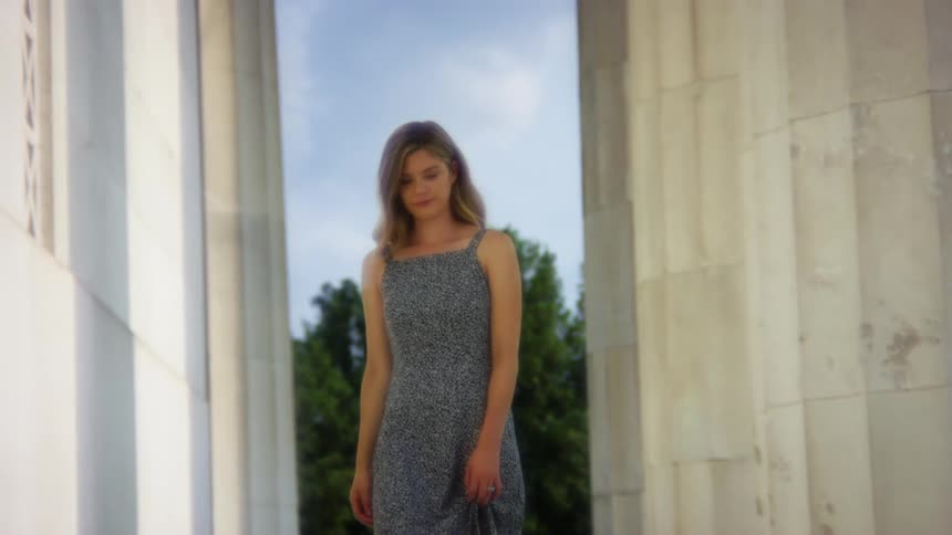A young woman slightly lifts her dress with one hand as she walks outside of a building on a sunny day