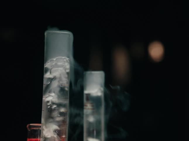 Two tall glass tubes bubble and steam near bottle with red liquid in dark area