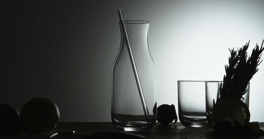 Fruit and vegetables sit near glass jug and cup in dim room briefly lit by light