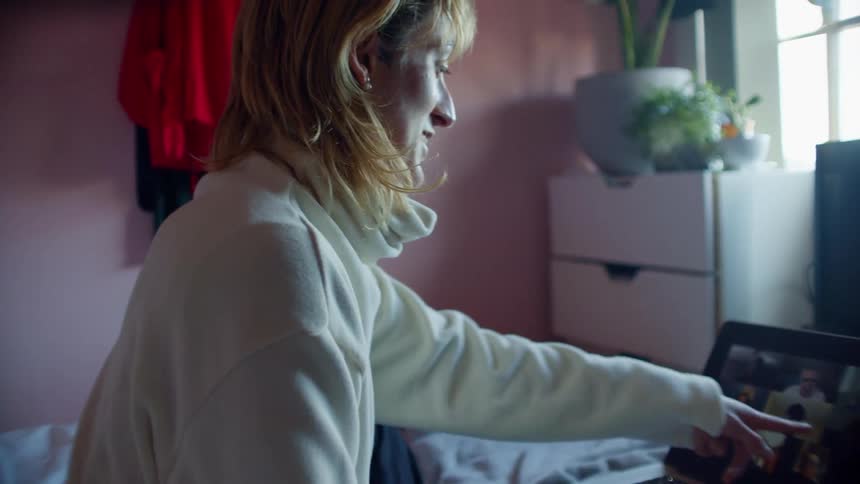 A young woman sitting on her bed points at her laptop as she video chats with people