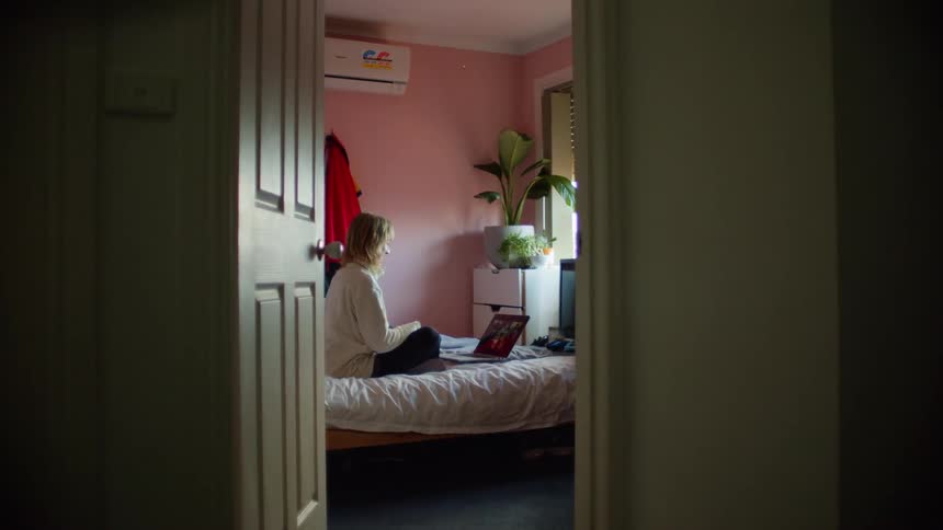 Blonde woman with a grey sweatshirt sitting on the bed makes a video call on the laptop and waves at the person