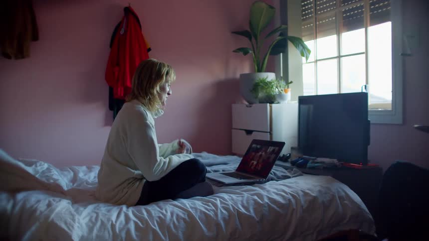 Blonde woman with a white turtleneck sweater sits on a bed and makes a video call on laptop in bedroom