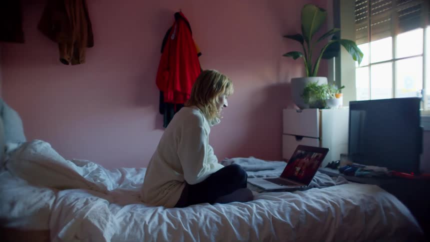 A woman uses a laptop to participate in a video chat while sitting on her bed