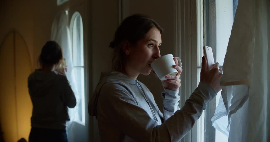 A woman stands by a window holding her cell phone up having a video call and takes a drink from a coffee mug