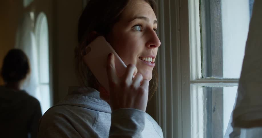 Woman with a grey hoodie talks on the smartphone smilingly while looking out the window