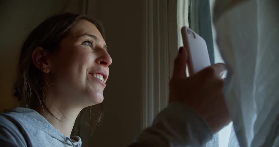 A young woman video chats on her phone while standing near a window