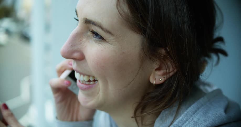 A young woman standing on a balcony with a coffee cup laughs as she speaks on her smartphone