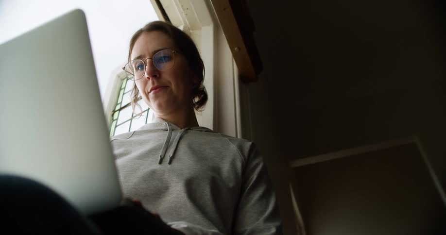 Woman sits in open near open window typing on laptop