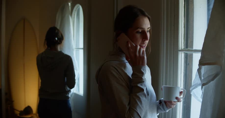 Woman with a grey hoodie holds a cup of coffee and talks on the smartphone smilingly while looking out the window