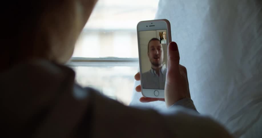 A woman uses a smartphone to video chat with a man as she stands near a window indoors during the day