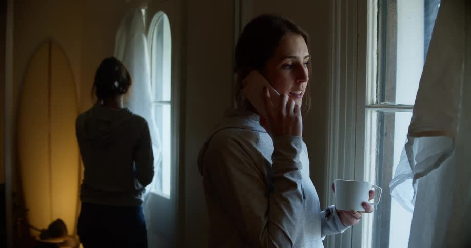 A young woman talks on her smartphone while standing near a window with a coffee mug