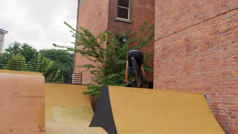 A female rides a small bicycle down a half pipe ramp that is set up between two large buildings