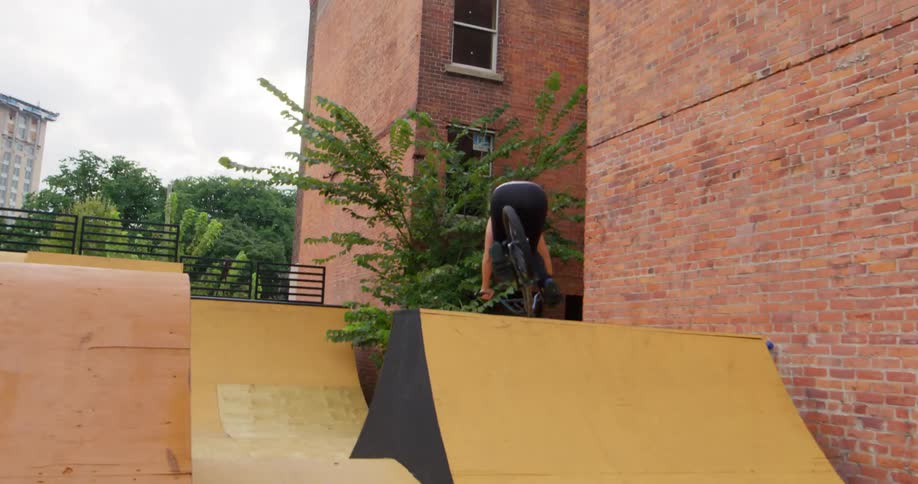 A female rides a small bicycle down a half pipe ramp that is set up between two large buildings