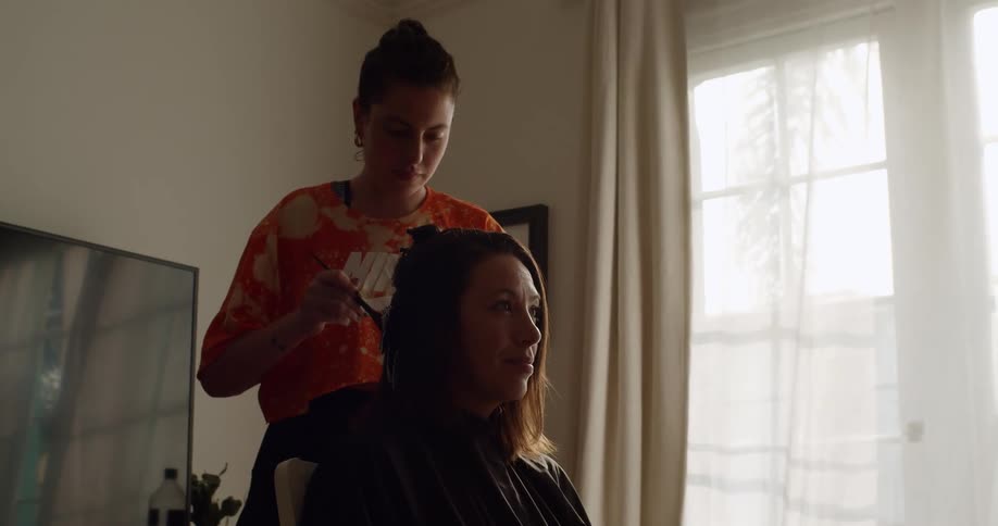 A woman stands behind a seated woman painting hair color into her hair