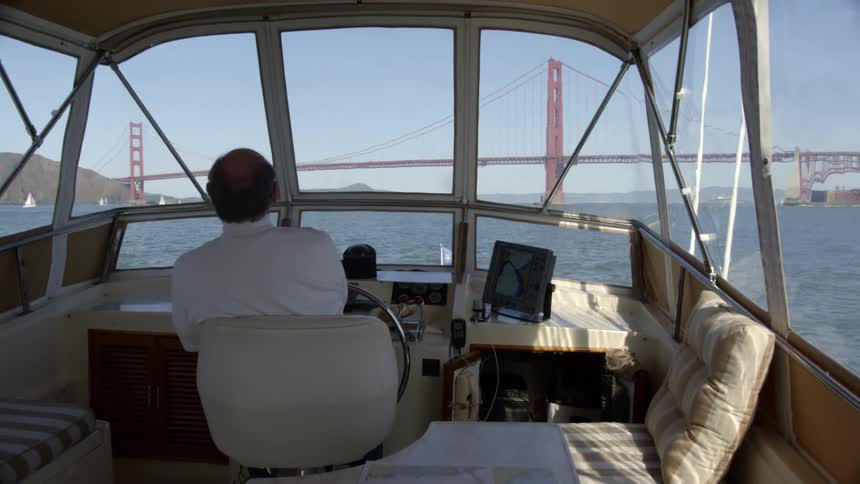 Man pilots boat toward suspension bridge while other boats float near shore