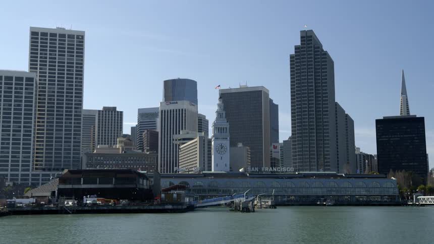 A clock tower with an american flag waving atop it on the coast of a metropolis on a sunny day