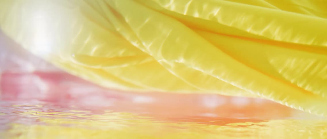 Yellow and purple fabric swirl and float in water as sunlight shimmers