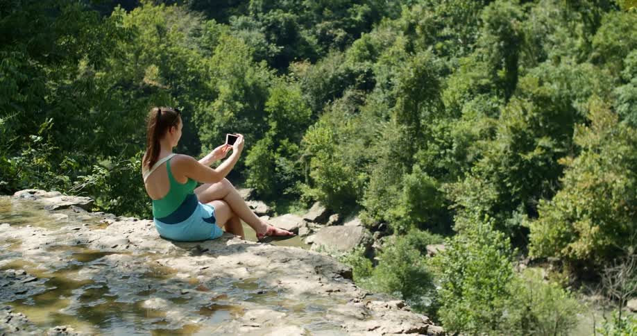 A teenage girl sitting on a cliff above a river takes a selfie on her cell phone