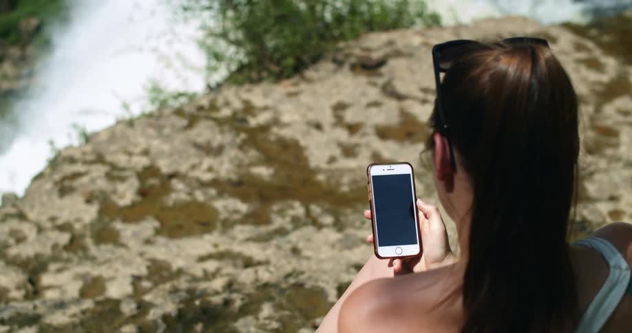 A woman with sunglasses next to a river and waterfall using a smart phone