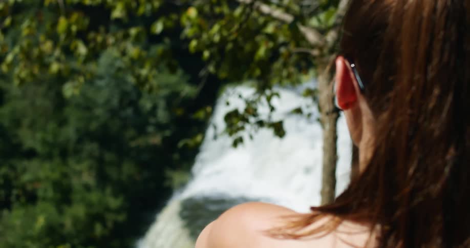 Woman wearing sunglasses looks toward waterfalls bordered by forest