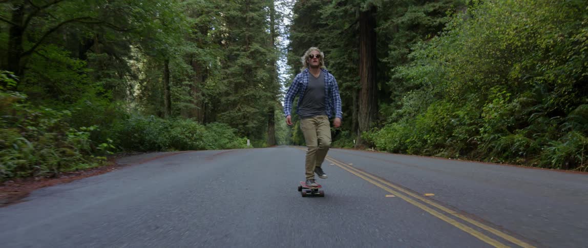 Blonde man riding his skateboard down a two-lane road through a wooded area