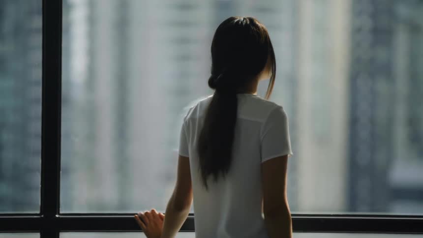 A young asian woman looks out her apartment window at the apartments across from her