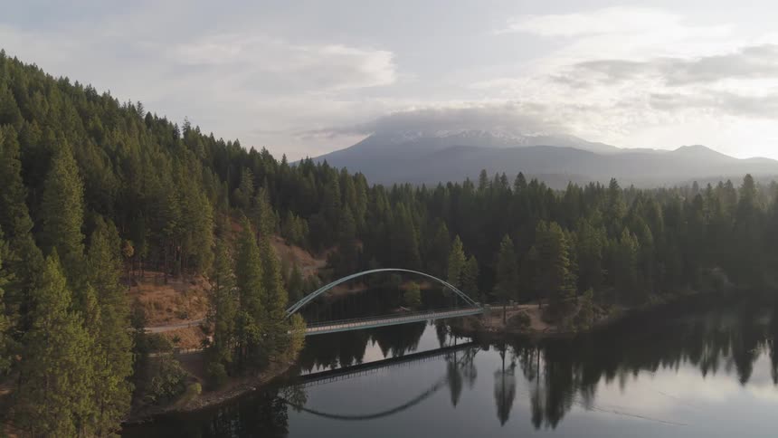 An aerial view of a bridge spanning a lake in a mountain forest in the daytime