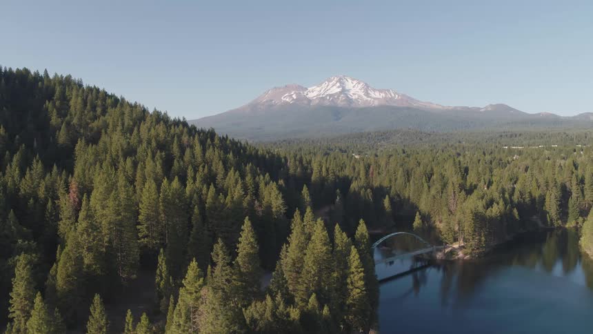 An aerial view of a forest and lake with a bridge with a mountain range in the distance