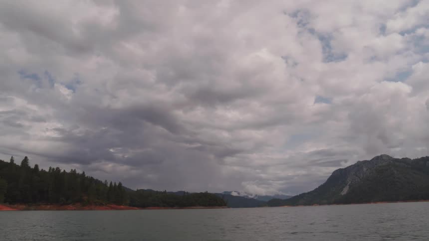 Conifers cover mountain bordering lake under clouds