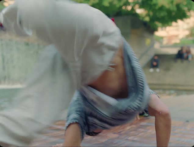 A teenage boy dances in front of a fountain in a public park as his friend watches and a group of people sitting on steps watch