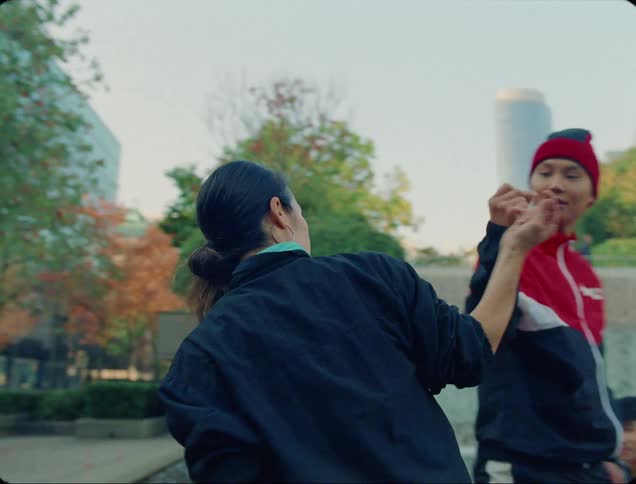A woman greets a man with an elaborate gesture before beginning to dance on a dance pad in front of a fountain in a city park during the day