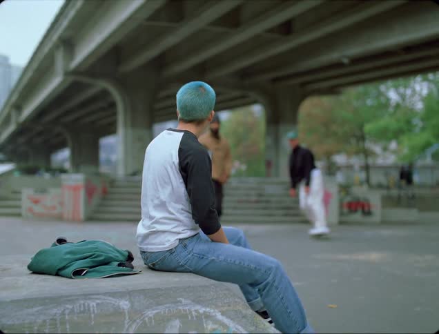 Two skateboarders perform jumps down stairs under an underpass near downtown skyscrapers and take time to high five a sitting skateboarder