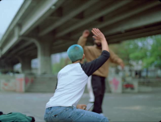 Young man with blue hair gives air high fives to skateboarders at skate park under overpass